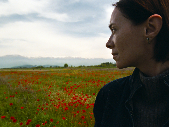 Une femme au profil déterminé regarde au loin, entourée d'un champ de coquelicots rouges éclatants, avec en arrière-plan des montagnes enneigées sous un ciel partiellement nuageux.