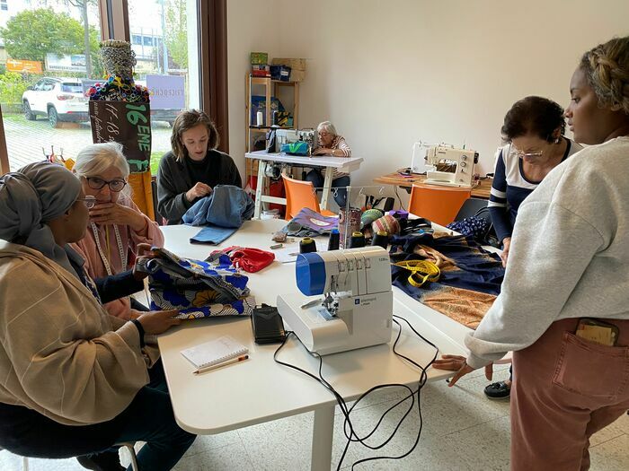 Sur cette photo, on aperçoit l'atelier de customisation de vêtements et accessoires. Cinq participants, aux origines et âges variés, travaillent autour d'une grande table, entourés de tissus, fils et outils. Ángela, l'animatrice, les guide dans la réparation et la création des vêtements qu'ils ont apportés. Chacun se concentre sur sa tâche : certains recousent, d'autres ajoutent des décorations ou ajustent des détails. L'ambiance est chaleureuse et collaborative, où l'échange entre cultures et…