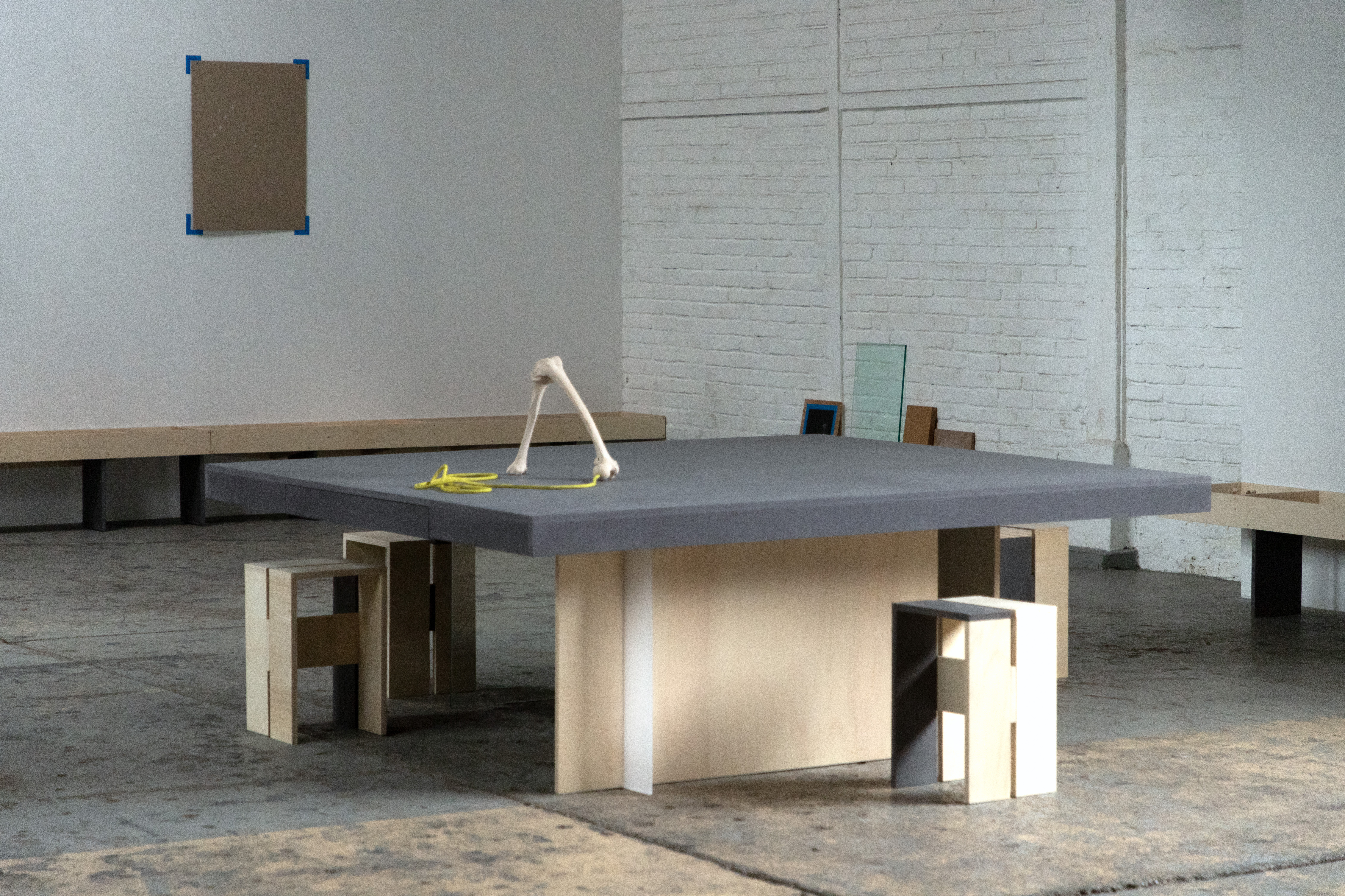 Une vue de l'installation Bones Score - une table en bois est entourée de 4 tabourets. Le plateau de la table est peint en gris. Sur la table un os et une cordelette jaune. Il n'y a pas de personnages sur cette image.