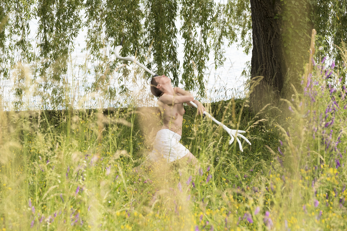 Une performer tenant une grande barre blanche qui se termine aux aux extrémités par des formes qui ressemblent à des pattes de poulet galope dans un champs. La performer porte un short blanc. Elle regarde en l'air. Il y a un grand arbre sur la droite de type saule pleureur. Ses branches touchent presque le sol. Les sol est très vert avec des herbes hautes.