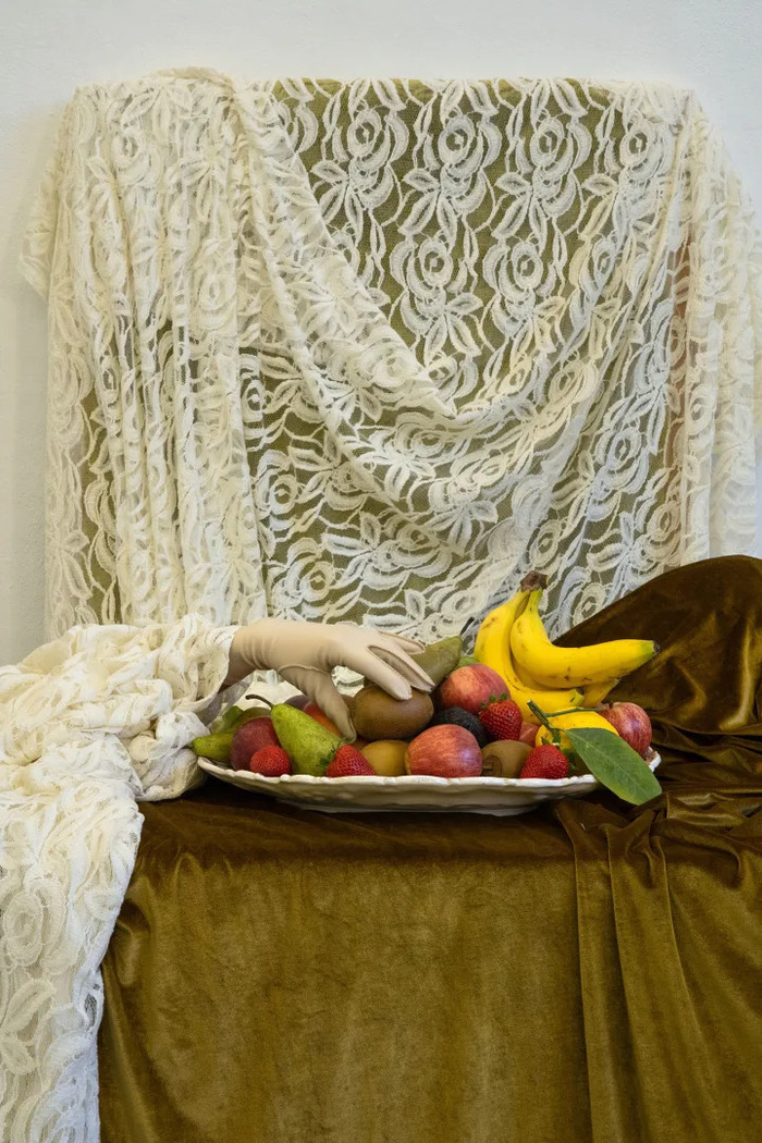 Un platêau de fruits est dressé sur une table nappée d'une nappe en velours beige. Une main gantée attrape un kiwi.