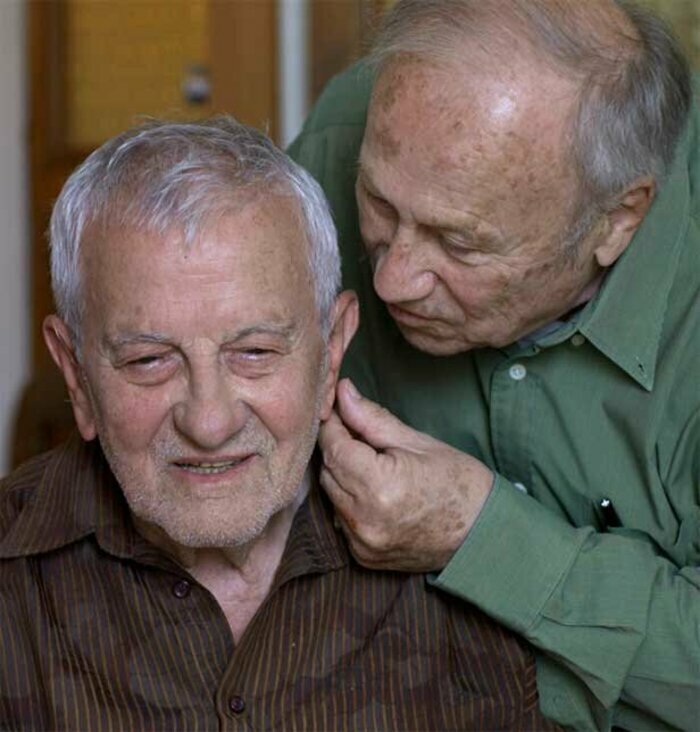 Deux homme âgés, proches l'un de l'autre, l'un des hommes dit un secret à l'oreille de l'autre homme.