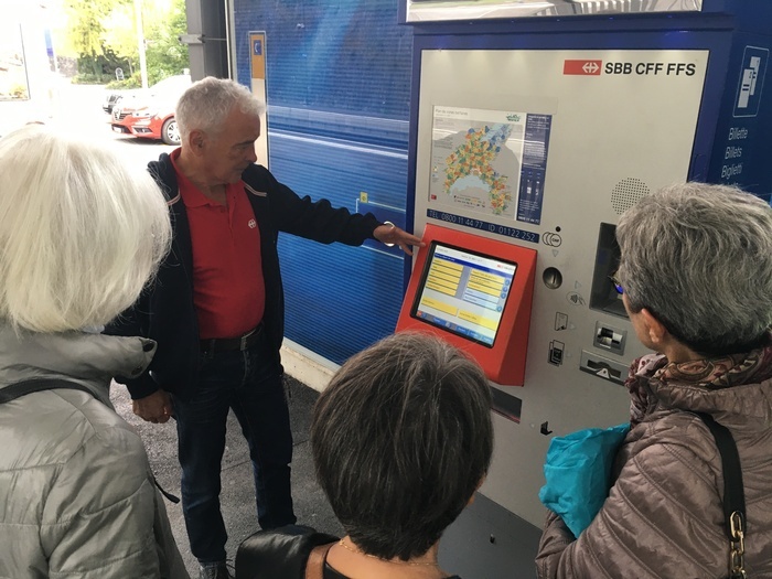 Plusieurs personnes autour d'une machine pour prendre un billet de train ; un homme semble expliquer comment utiliser la machine pour acheter un billet.