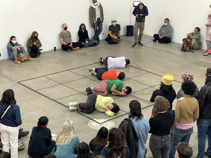 Dans une salle d'exposition, quatre performers sont couché·e·s au sol sur un échiquier de 4 carreaux sur 4 carreaux, un public entoure la scène.