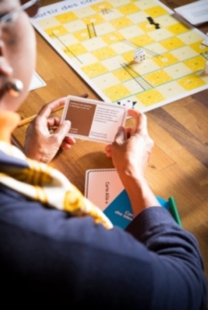 Sur cette image on voit une dame jouer à un jeu de société. Sur la table il y a un jeu avec un dès, et la dame est en trainde lire une carte qu'elle a entre les mains.