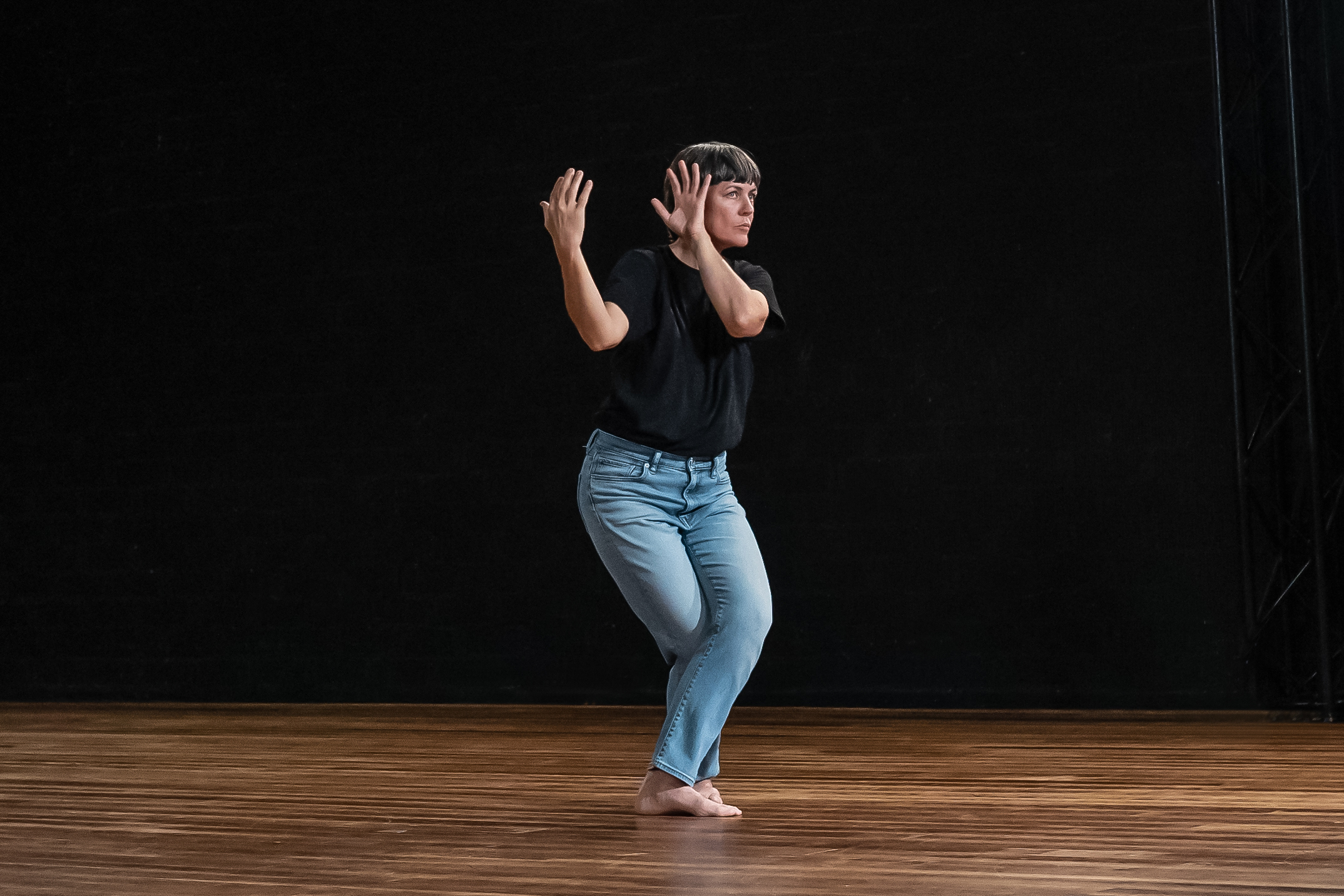 La danseuse DD Dorviliier se trouve au milieu d'une scène. Le sol est en bois, la lumière est diffuse. La danseuse est habillée d'un blue jeans et d'un t-shirt noir. Ses mains se trouvent à la hauteur de son visage sur la gauche et semblent tenir une sphère (absente). L'espacement entre les mains est d'environ 50 cm. La danseuse regarde en face d'elle.