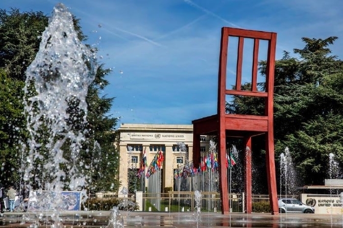 Jet d'eau avec immense chaise en bois. Au loin, un grand bâtiment dont l'entrée est bordée de drapeaux