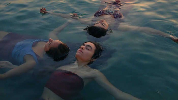 Trois femmes flottant paisiblement à la surface de l'eau, les yeux fermés, exprimant la sérénité, la résilience et l'abandon en pleine nature.