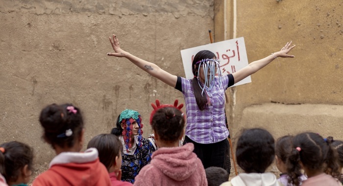 Une femme masquée aux bras écartés anime un spectacle de rue pour des enfants devant un mur en terre, accompagnée d'une autre personne en bonnet coloré.