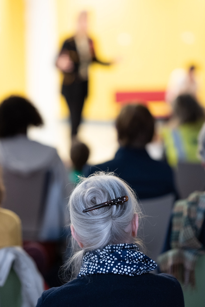 Une assemblée de personnes assises est photographiées de dos. La personne au premier plan est nette, les autres en arrière plan sont floues.