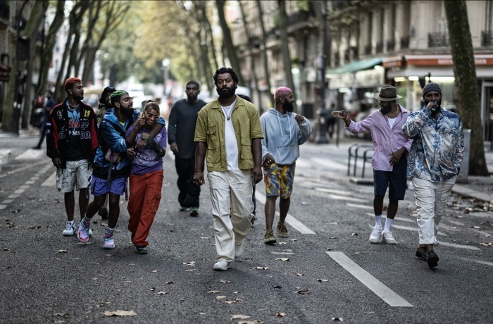 Un groupe d'amis déambule dans une rue bordée d'arbres, mêlant styles vestimentaires colorés et expressions décontractées. Au centre, un homme en chemise jaune marche avec assurance, entouré de rires et d'énergie. L'ambiance est urbaine, vibrante et pleine de vie.