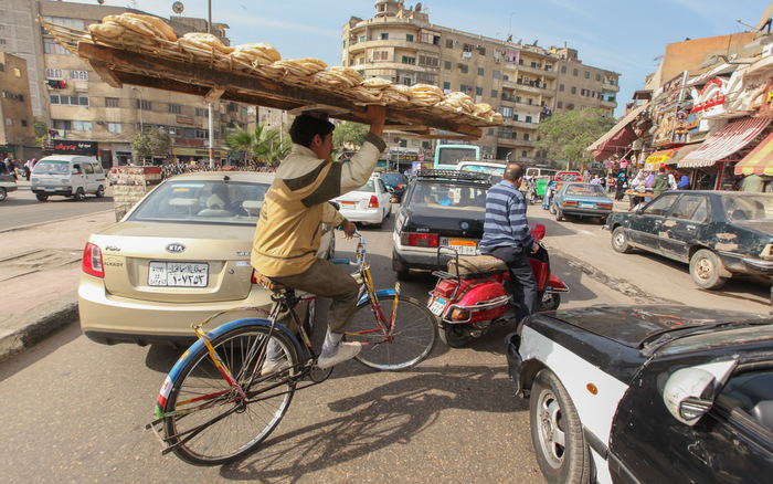 homme portant un plateau sur sa tête avec du pain, sur un vélo dans les rues du Caire, en Egypte - Claude Marthaler