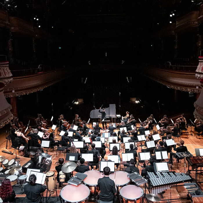 Nous voyons une grande scène de concert vue de haut. Un orchestre symphonique est installé en demi-cercle autour d’un chef d’orchestre debout, les bras levés, au centre de la scène. Les musiciens, en tenue formelle noire, jouent de divers instruments: des cordes (violons, altos, violoncelles), des bois, des cuivres et des percussions.

Au premier plan, on remarque les percussions: des timbales, des tambours et un xylophone. Le piano est visible au centre, en avant des musiciens. La salle est…