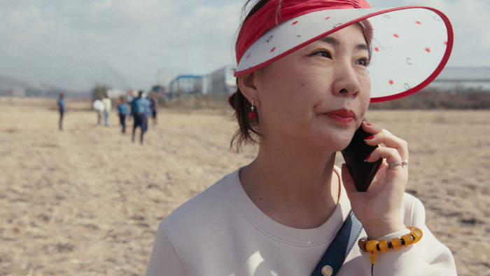 Une femme portant une visière rouge et blanche ornée d'étoiles, en communication téléphonique, se tient dans un champ avec des personnes floues en arrière-plan.