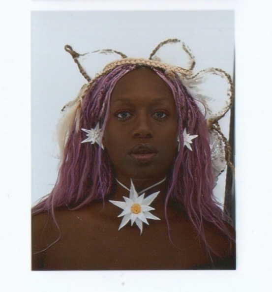 Portrait photographique d'une personne avec de longs cheveux violets tressés et des boucles d'oreille et un collier en forme d'edelweiss.