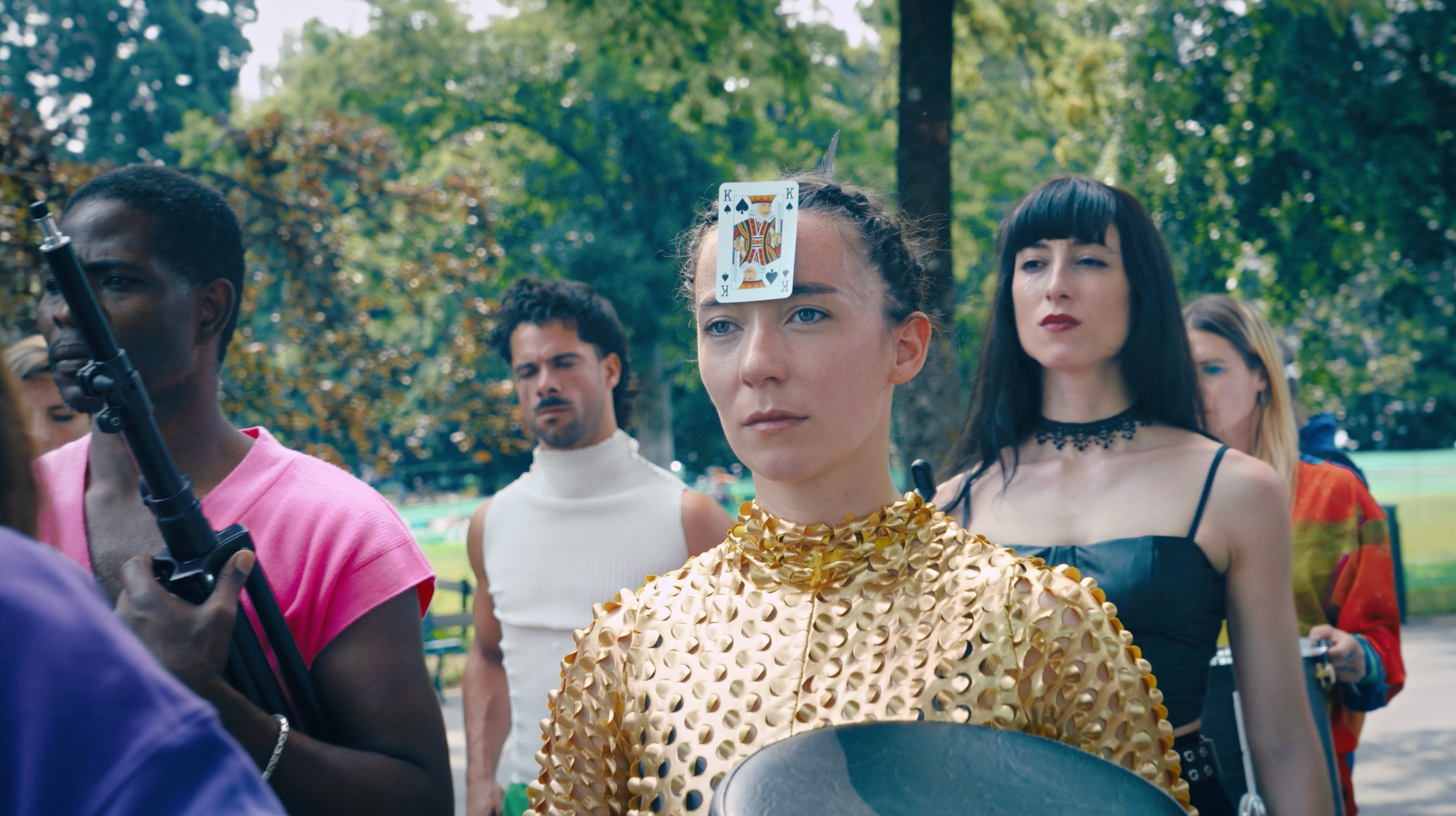 Un groupe de personnes dans un endroit verdoyant. Au centre de l'image, une performer portant un vêtement doré regarde hors cadre. Sur son front, une carte à jouer, celle du roi de pic.