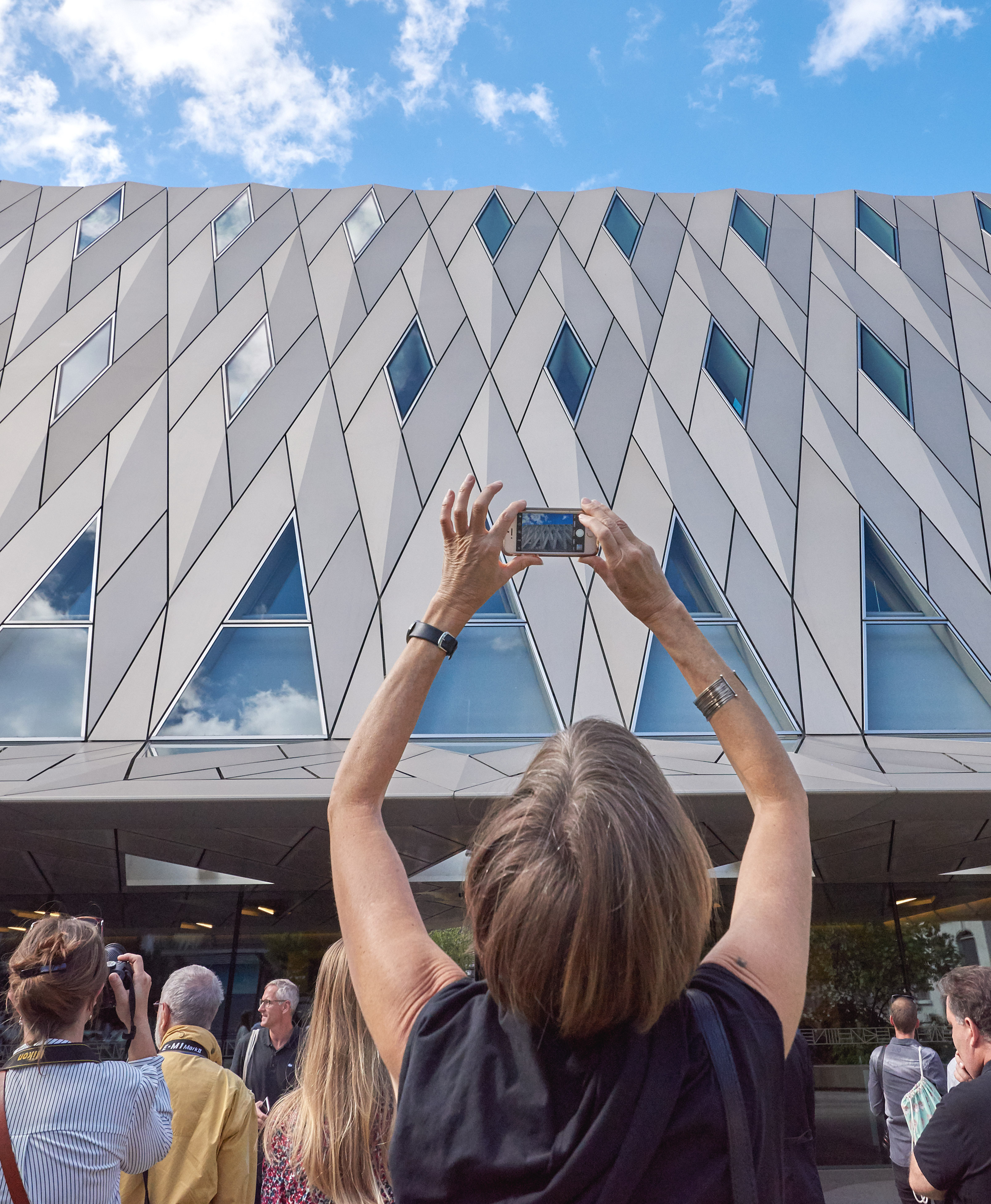 Une personne photographie le bâtiment du MEG avec son portable. Elle est de dos avec d'autres personnes.