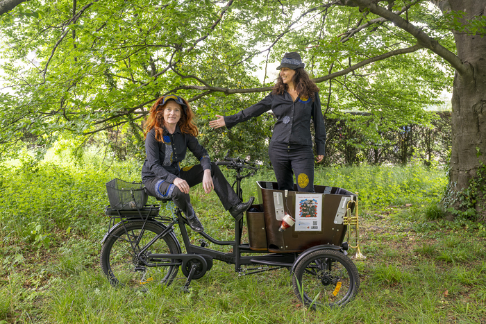 Deux dames habillé en noir qui se trouve sur un vélo à 3 roues qui possède un chariot à l'avant. Elles sont dans une forêt, à l'arrière plan on voit un grand arbre à droite et des buissons plus loin.