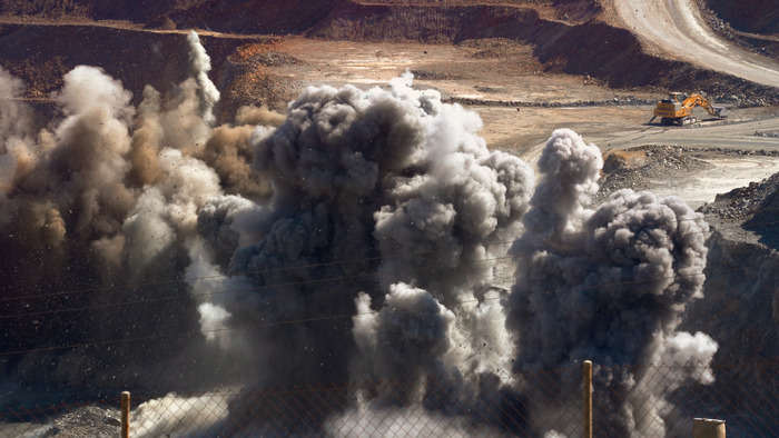 Une explosion massive dans une mine à ciel ouvert projette d'épais nuages de fumée grise dans les airs. En arrière-plan, une pelleteuse jaune.