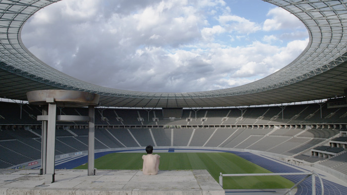 Il s'agit d'un still du film dans lequel ou voit un stade de football vide. Au premier plan est assise une personne, dos à nous qui regarde l'intérieur du stade. Elle est nue.