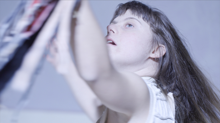 Une jeune personne danse dans un espace très éclairé. Elle tient un tissu à la main (flou) qu'elle regarde intensément.