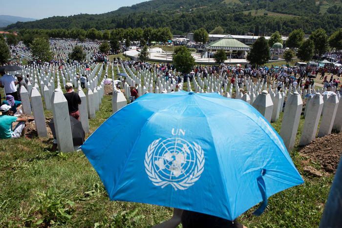 Une grande foule se rassemble dans le cimetière mémorial de Srebrenica, entourée de rangées de stèles blanches symbolisant les victimes du massacre de 1995. Au premier plan, un parapluie bleu portant le logo des Nations Unies (UN). Le paysage verdoyant et les montagnes en arrière-plan.