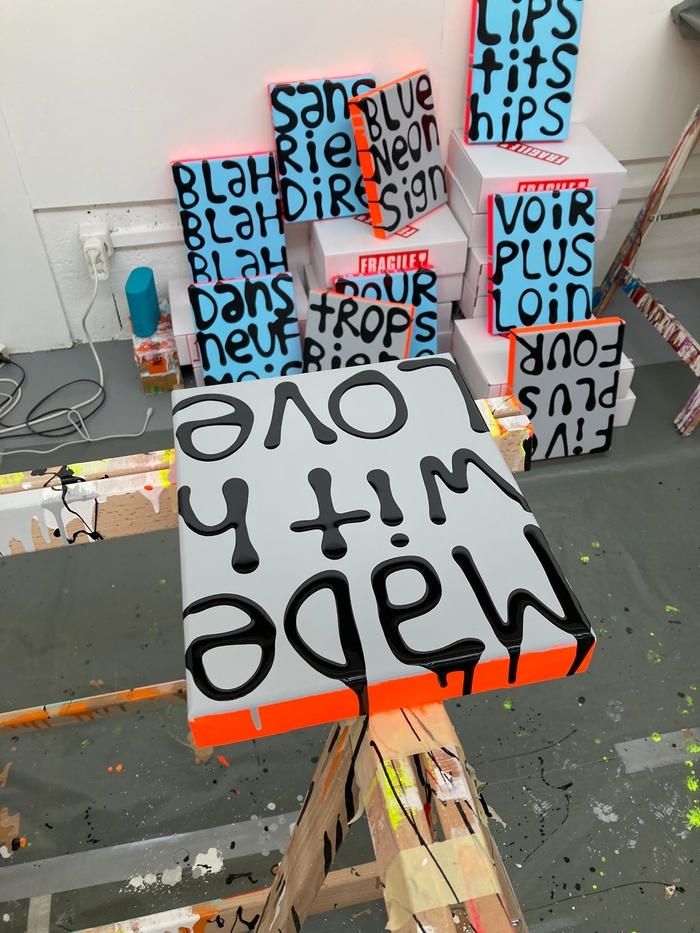 Nous sommes dans l'atelier de l'artiste Hadrien Dussoix. L'image montre plusieurs tableaux colorés, sur lesquels apparaissent des textes brefs écrits en grosses lettres, en peinture acrylique.