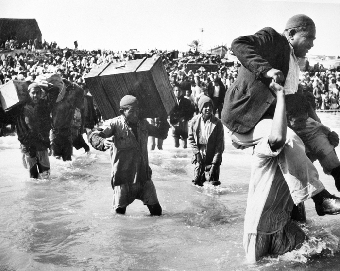 Image en noir et blanc montrant des réfugiés portant des valises et traversant l'eau, entourés d'une foule dense, illustrant l'exode et les défis humanitaires.