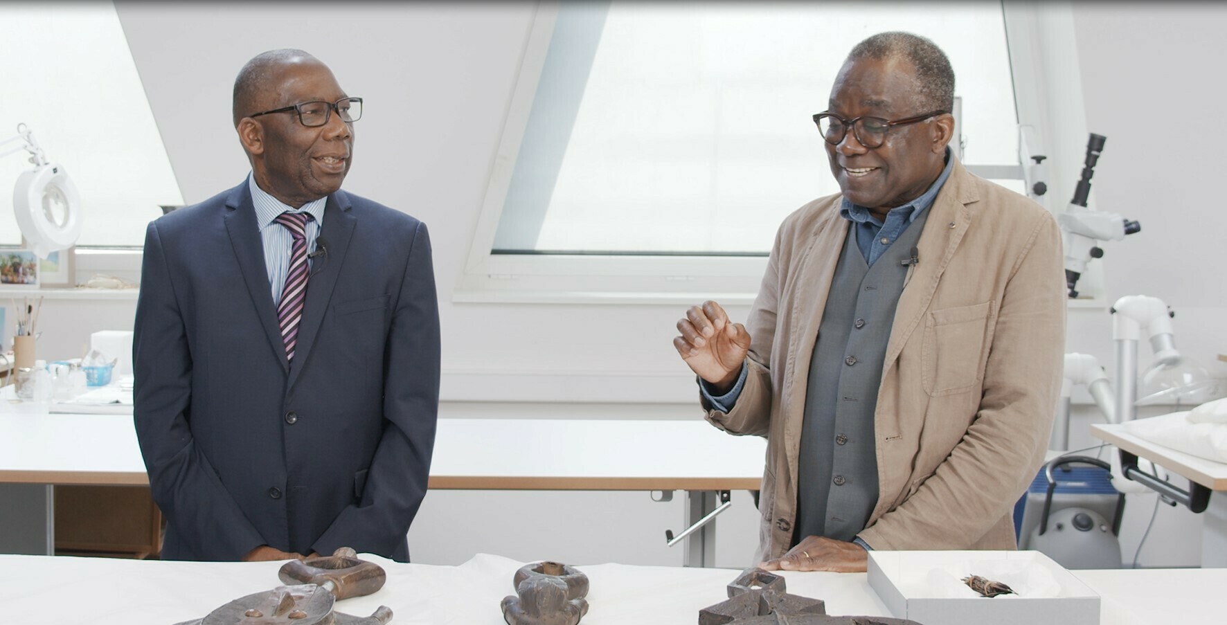 Jean Lignongo et Jean-Didier Ekori sont face caméra, devant une table sur laquelle on devine des statues de reliquaires Kota. L'un parle et sourit, l'autre le regarde.