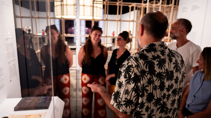 Visite guidée dans l'exposition temporaire. Le guide est de dos et interagit avec un groupe de 5 personnes. Les participant-e-s sont souriant et l'ambiance est chaleureuse.