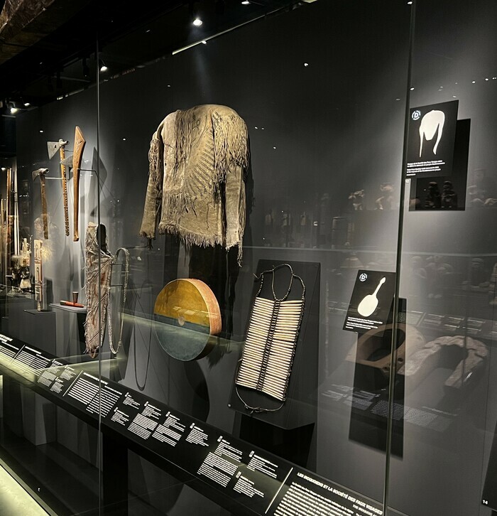 Photographie d'une vitrine de la section Amériques de l'exposition permanente du MEG. Deux objets de cette vitrine, appartenant au peuple autochtone Haudenosaunee, ont été restitués en février 2023. Une silhouette de l'objet découpée dans un carré les remplace.