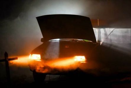 Une voiture avec une porte et le capot ouvert ainsi que les feux allumés dans l'obscurité.