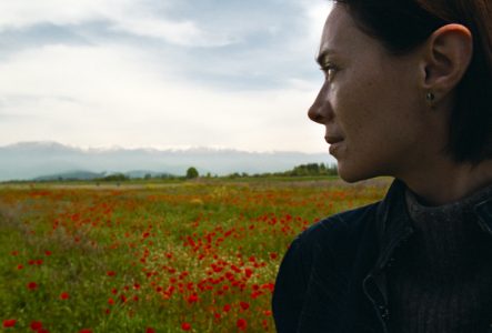 Une femme au profil déterminé regarde au loin, entourée d'un champ de coquelicots rouges éclatants, avec en arrière-plan des montagnes enneigées sous un ciel partiellement nuageux.