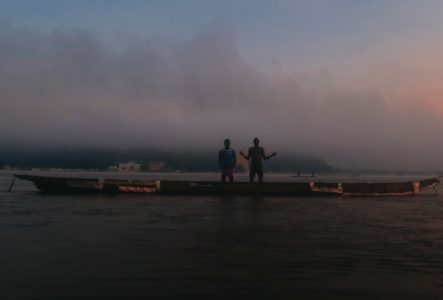 Deux personnes debout sur une barge au crépuscule, entourées de brume, avec des bâtiments en arrière-plan, évoquant un moment de calme ou de réflexion sur les rives d'une ville africaine.