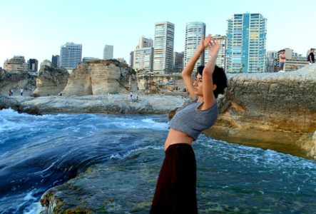 Jeune femme levant les bras face à la mer agitée à Beyrouth, avec en arrière-plan des falaises emblématiques et des immeubles modernes.