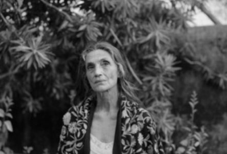 Une femme pensive, vêtue de fleurs, se tient au milieu d'un jardin sauvage. Son regard mélancolique flotte dans le vide.