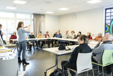 Une dame réalise une présentation face à des seniors attablées, qui l'écoutent attentivement.