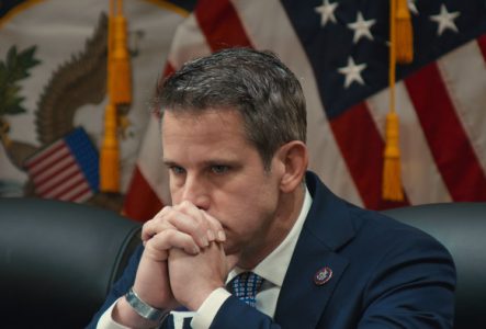 Adam Kinzinger en costume, l'air pensif et préoccupé, assis devant des drapeaux américains. Ses mains jointes devant son visage traduisent une intense concentration ou réflexion.