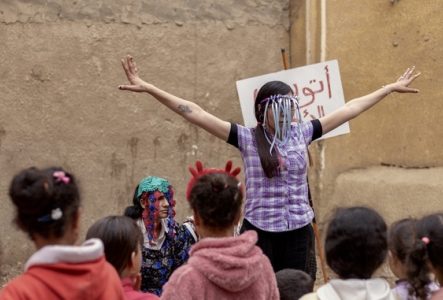 Une femme masquée aux bras écartés anime un spectacle de rue pour des enfants devant un mur en terre, accompagnée d'une autre personne en bonnet coloré.