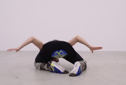 Une personne portant des pantalons moulants de sport est à genoux sur le sol d'un "white cube". Les bras sont repliés sur les côtés comme les ailes d'une mouette. La personne est positionnée les pieds face à nous et la tête en direction mur.