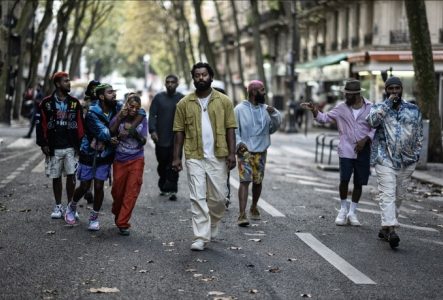 Un groupe d'amis déambule dans une rue bordée d'arbres, mêlant styles vestimentaires colorés et expressions décontractées. Au centre, un homme en chemise jaune marche avec assurance, entouré de rires et d'énergie. L'ambiance est urbaine, vibrante et pleine de vie.