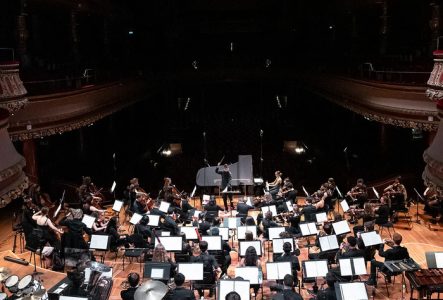 Nous voyons une grande scène de concert vue de haut. Un orchestre symphonique est installé en demi-cercle autour d’un chef d’orchestre debout, les bras levés, au centre de la scène. Les musiciens, en tenue formelle noire, jouent de divers instruments: des cordes (violons, altos, violoncelles), des bois, des cuivres et des percussions.

Au premier plan, on remarque les percussions: des timbales, des tambours et un xylophone. Le piano est visible au centre, en avant des musiciens. La salle est…