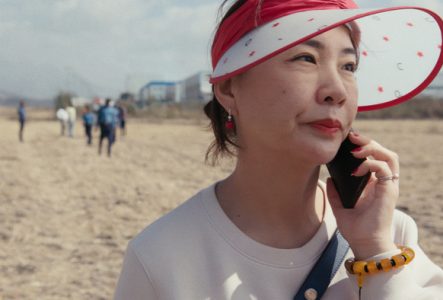 Une femme portant une visière rouge et blanche ornée d'étoiles, en communication téléphonique, se tient dans un champ avec des personnes floues en arrière-plan.