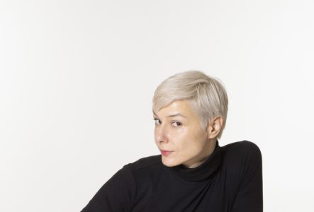 Une femme blanches aux cheveux courts avec un pull noir pose devant la caméra. C'est l'écrivaine Ed Wige.