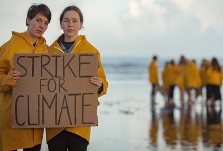 Deux jeunes personnes en cirés jaunes tenant une pancarte "STRIKE FOR CLIMATE" sur une plage, avec un groupe en arrière-plan, symbolisant l'engagement des jeunes pour la justice climatique.