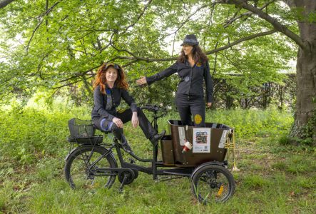 Deux dames habillé en noir qui se trouve sur un vélo à 3 roues qui possède un chariot à l'avant. Elles sont dans une forêt, à l'arrière plan on voit un grand arbre à droite et des buissons plus loin.