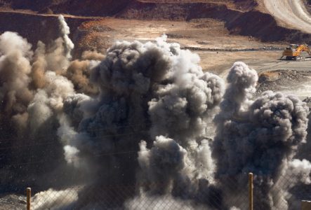 Une explosion massive dans une mine à ciel ouvert projette d'épais nuages de fumée grise dans les airs. En arrière-plan, une pelleteuse jaune.