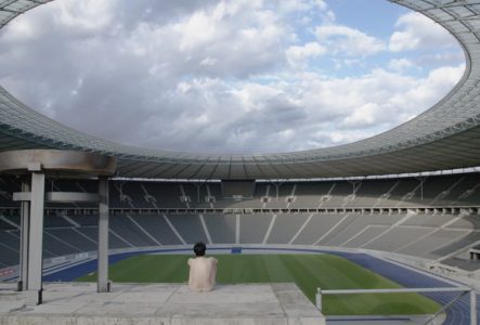 Il s'agit d'un still du film dans lequel ou voit un stade de football vide. Au premier plan est assise une personne, dos à nous qui regarde l'intérieur du stade. Elle est nue.