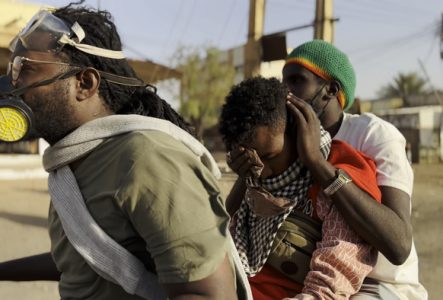 Un homme portant un masque à gaz conduit un véhicule avec deux passagers, dont un enfant protégé par un adulte portant un bonnet coloré, illustrant la résilience et la protection en temps de crise au Soudan.