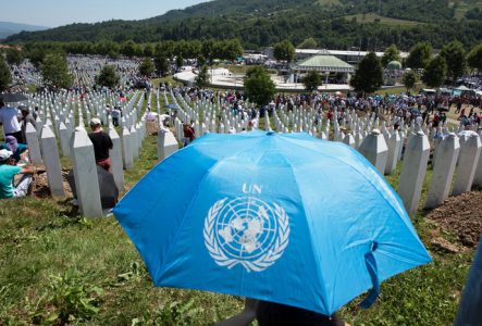 Une grande foule se rassemble dans le cimetière mémorial de Srebrenica, entourée de rangées de stèles blanches symbolisant les victimes du massacre de 1995. Au premier plan, un parapluie bleu portant le logo des Nations Unies (UN). Le paysage verdoyant et les montagnes en arrière-plan.